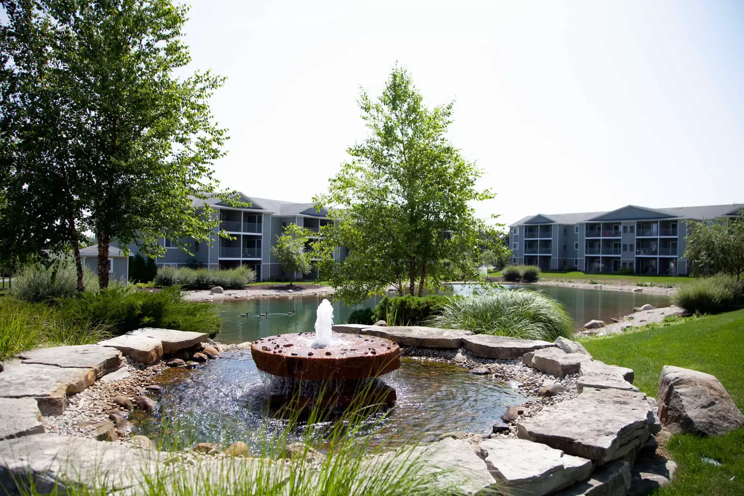 A fountain circled by rocks flows into the pond in front of the apartment buildings.